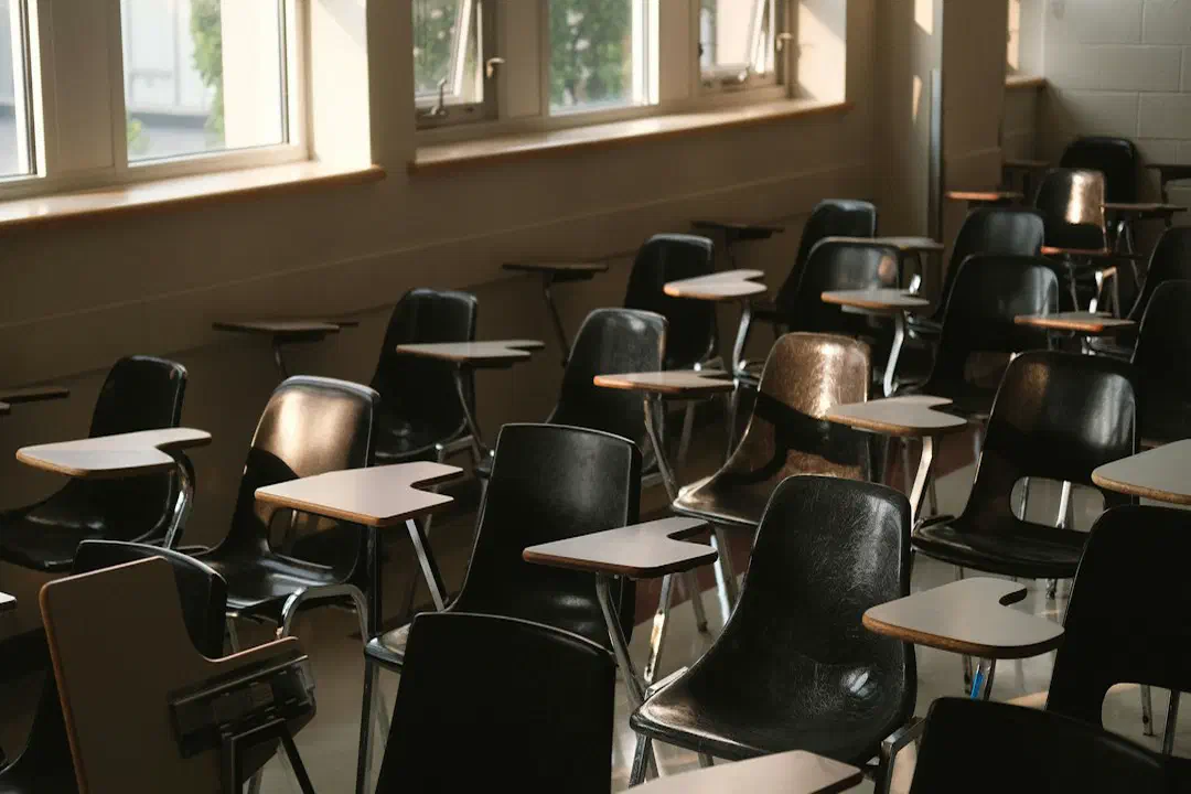 An empty classroom with rows of wooden desks and chairs beside sunlit windows
