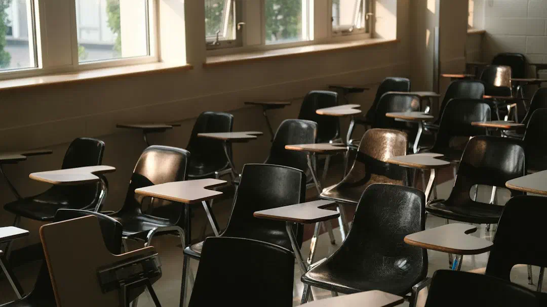 An empty classroom with rows of wooden desks and chairs beside sunlit windows